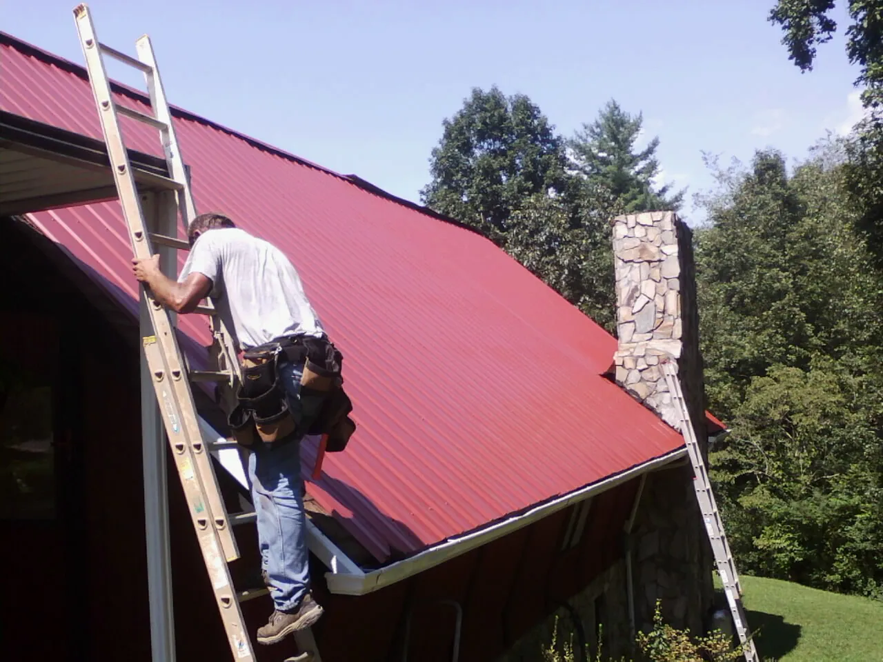 Roofer on a ladder beside a red metal roof and stone chimney — Soffit & Fascia Repair by Roofing Brothers in Beech Bottom